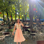 A woman wearing a traditional German Dirndl dress Berwin Wolf with a floral bodice and a light pink skirt stands in an outdoor Beergarden