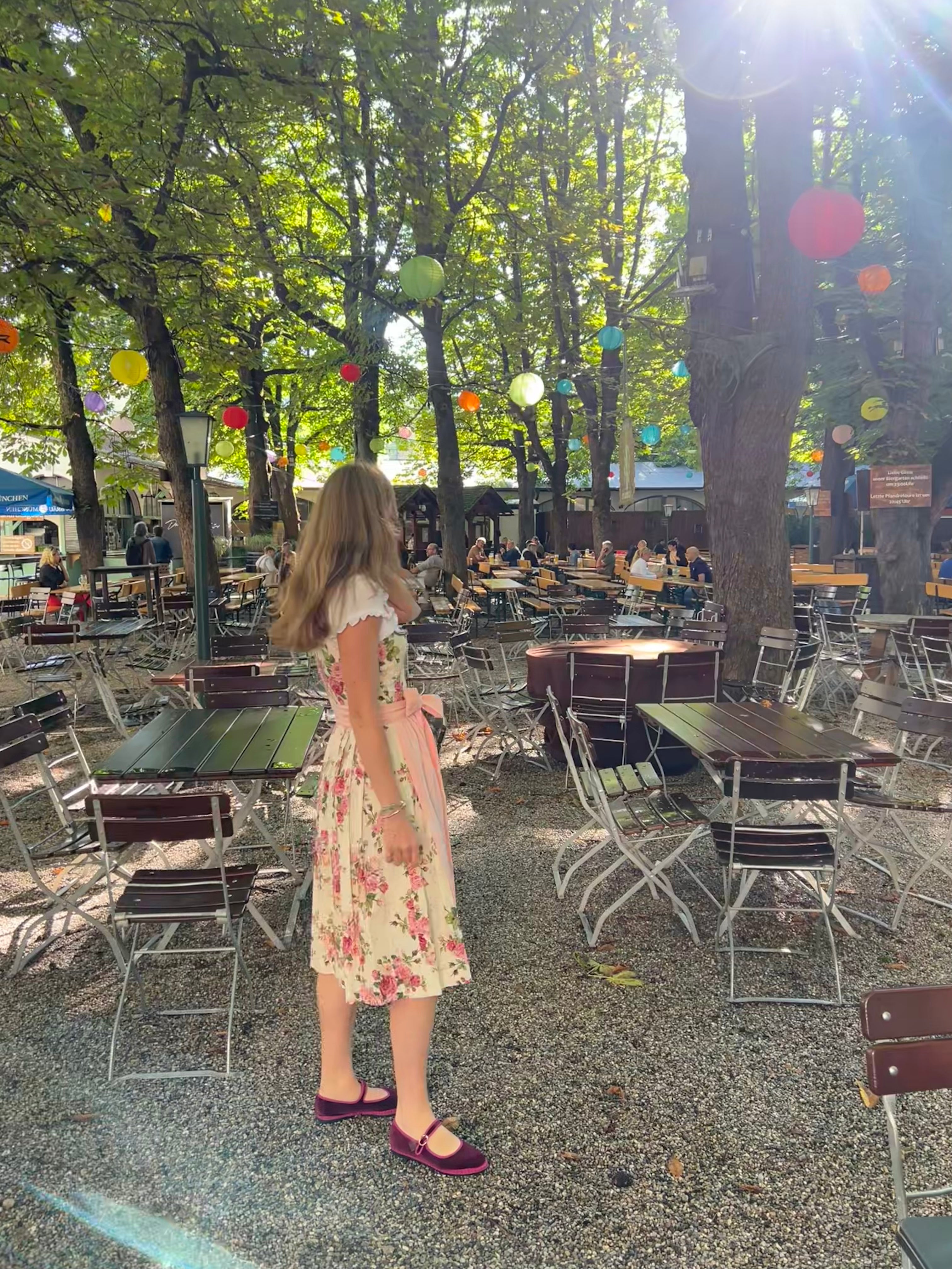A woman wearing a traditional German Dirndl dress Berwin Wolf with a floral bodice and a light pink skirt stands in an outdoor Beergarden