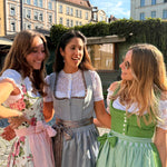Three women wearing traditional German dirndl dresses stand together outdoors in a sunny town square. The dress on the left is white with pink floral patterns and a pink apron, the center dress is a dusty blue-gray with brown suspenders, and the dress on the right is green and white checkered with a white bodice.