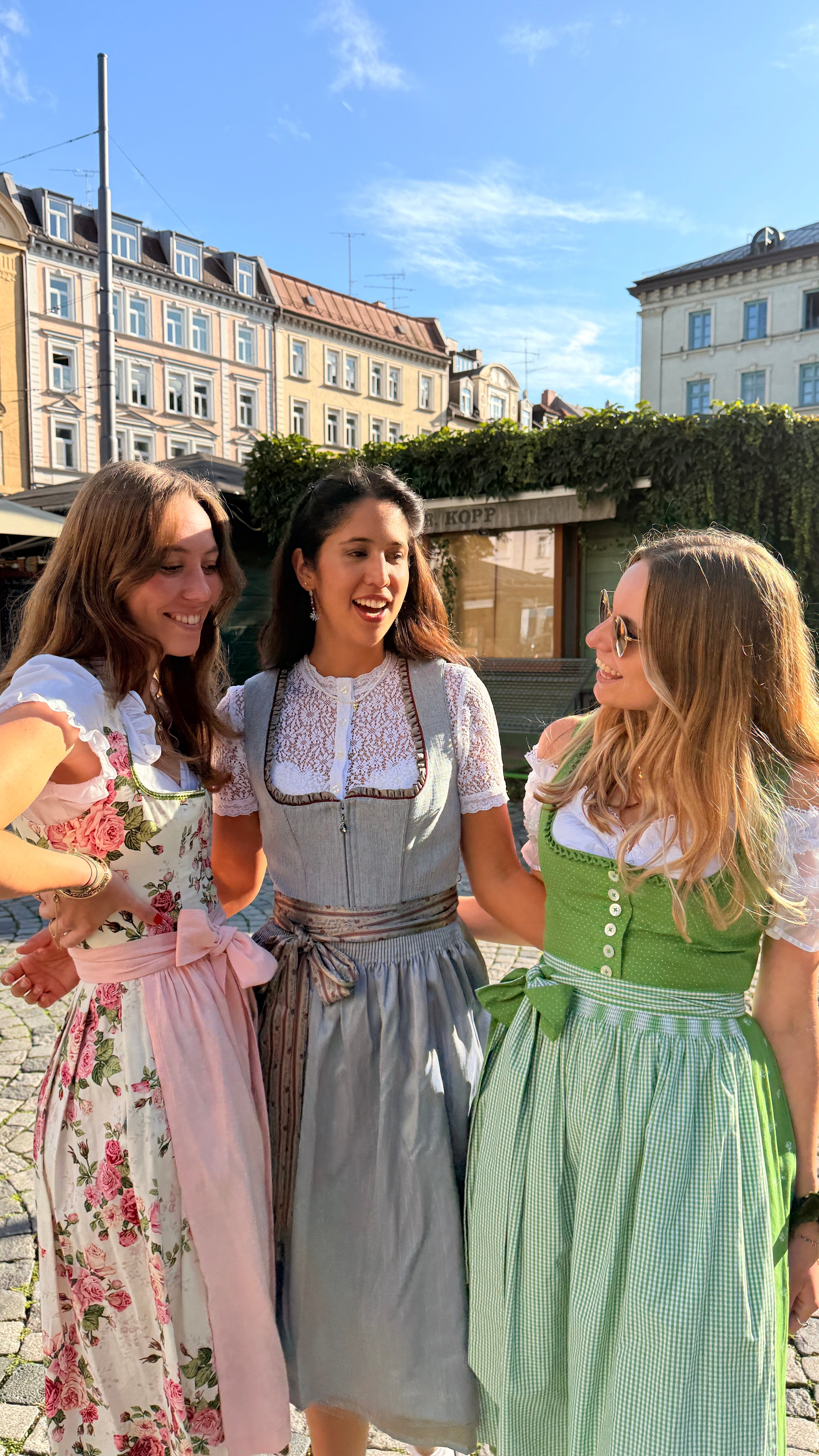Three women wearing traditional German dirndl dresses stand together outdoors in a sunny town square. The dress on the left is white with pink floral patterns and a pink apron, the center dress is a dusty blue-gray with brown suspenders, and the dress on the right is green and white checkered with a white bodice.