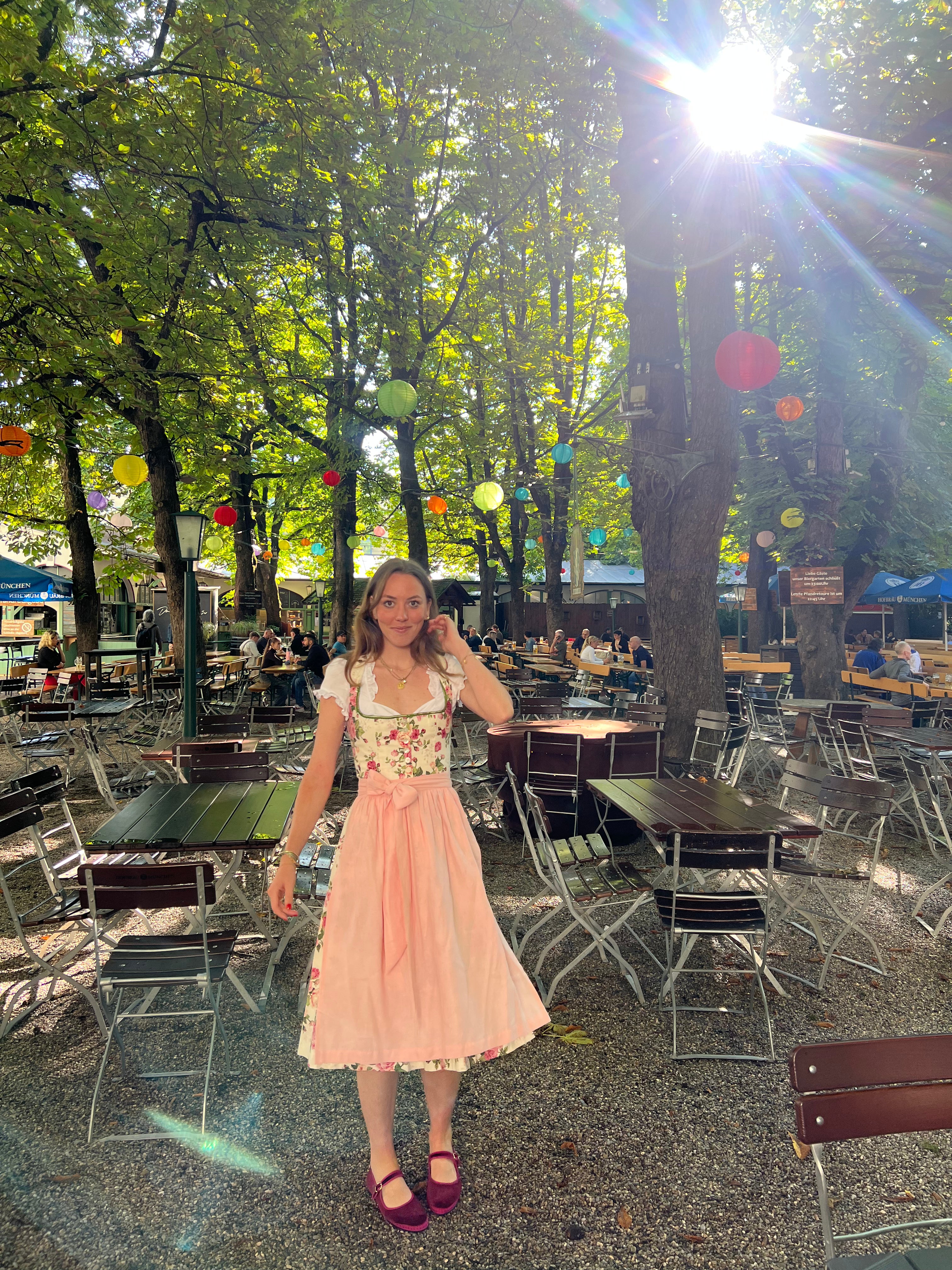 A woman wearing a traditional German Dirndl dress Berwin Wolf with a floral bodice and a light pink skirt stands in an outdoor Beergarden