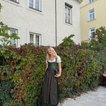 Woman in traditional green and brown Berwin and Wolf German dirndl standing in front of a house with greenery, ready for Oktoberfest 