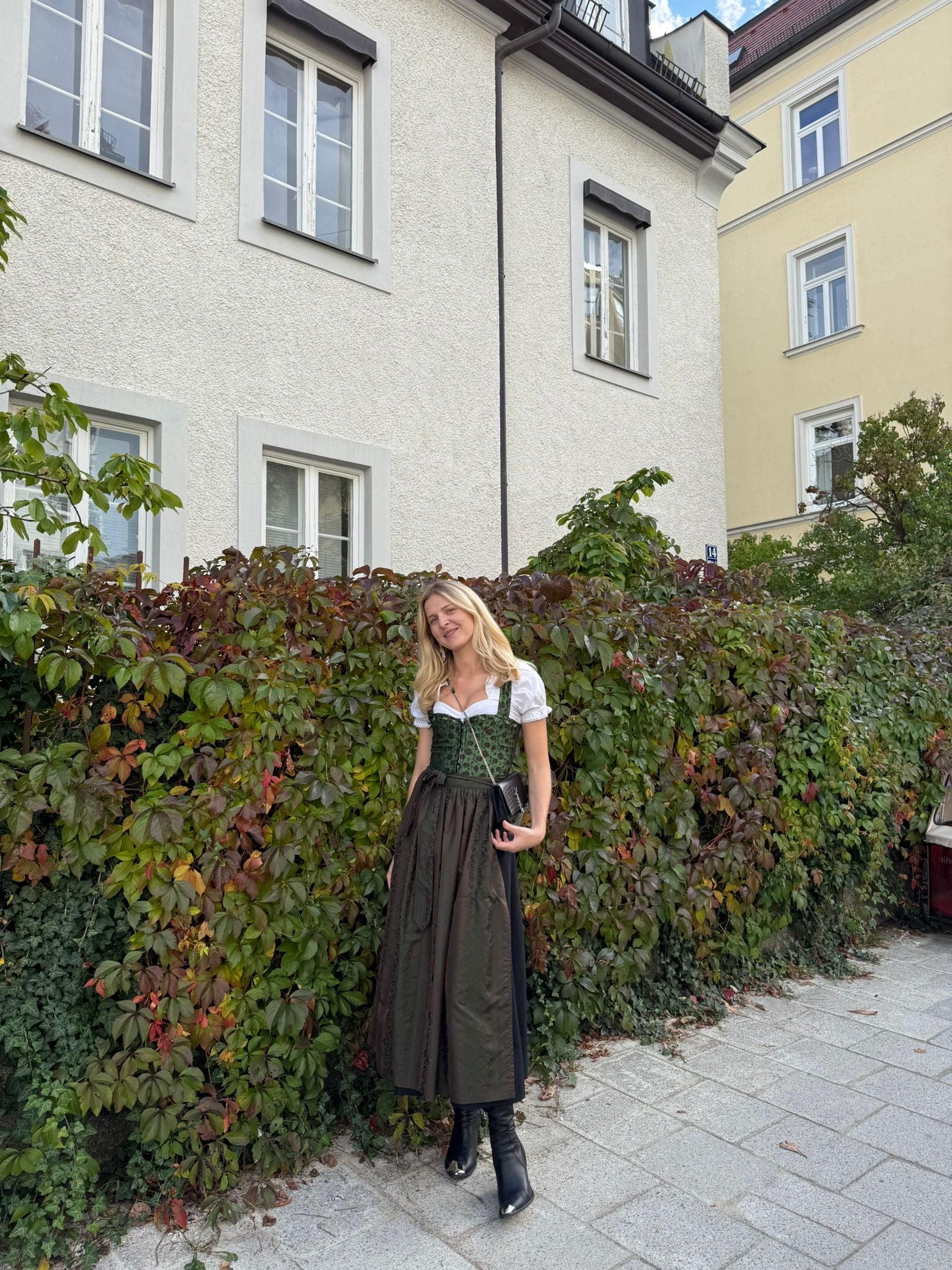 Woman in traditional green and brown Berwin and Wolf German dirndl standing in front of a house with greenery, ready for Oktoberfest 