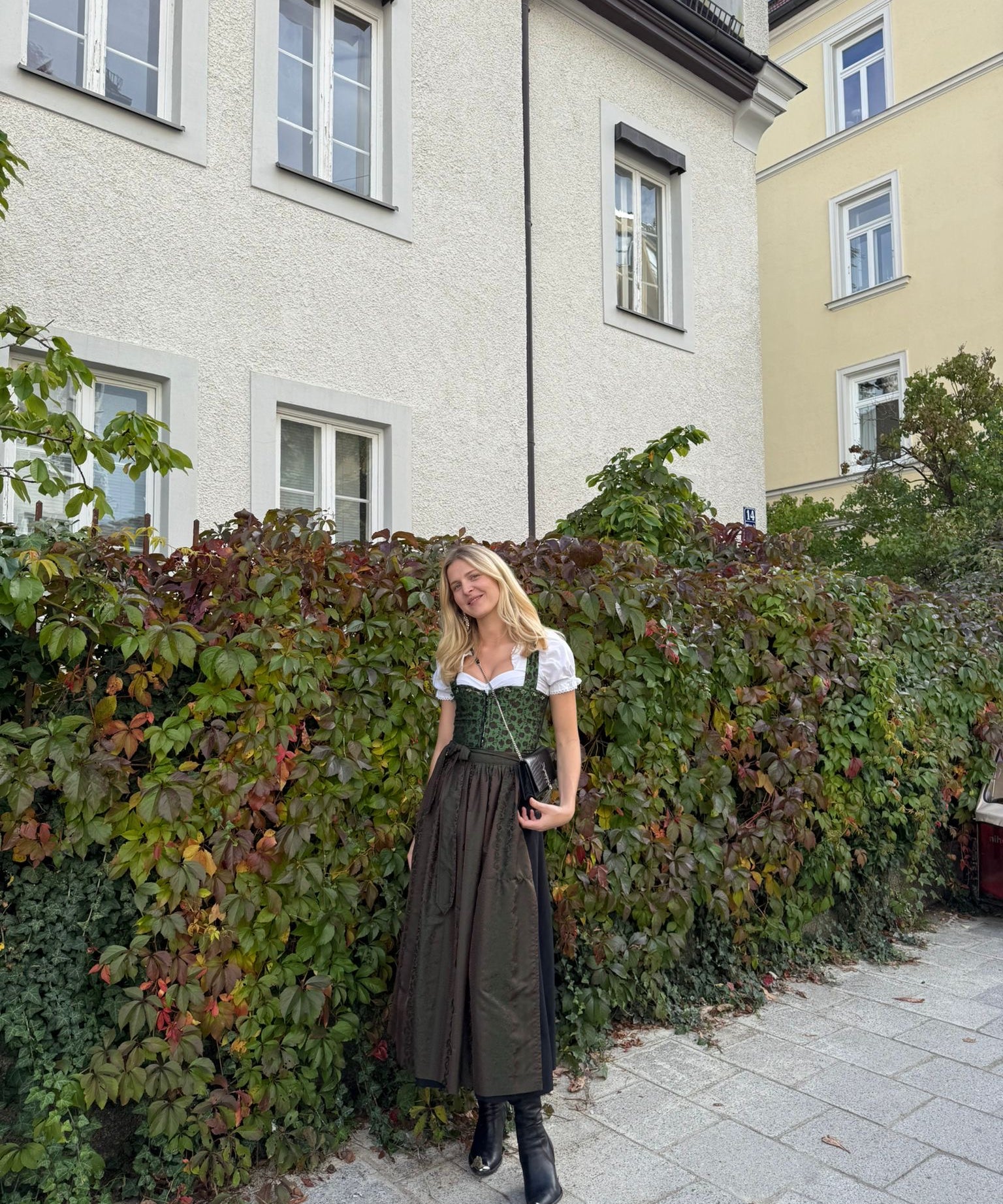 Woman in traditional green and brown Berwin and Wolf German dirndl standing in front of a house with greenery, ready for Oktoberfest 