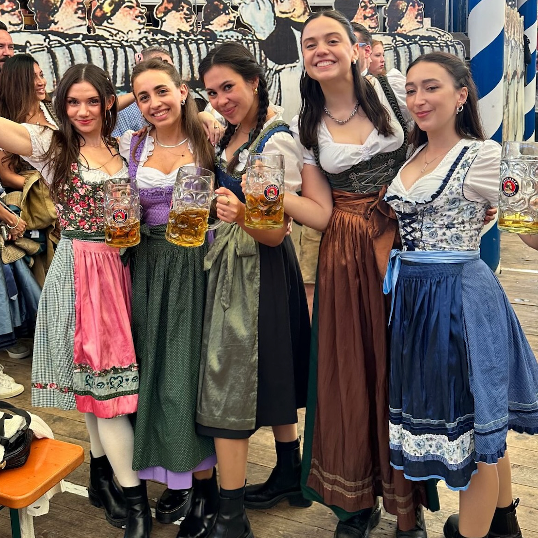 Five women in traditional German dirndl outfits posing together and holding large beer steins on a wooden deck in front of a Bavarian-themed backdrop.