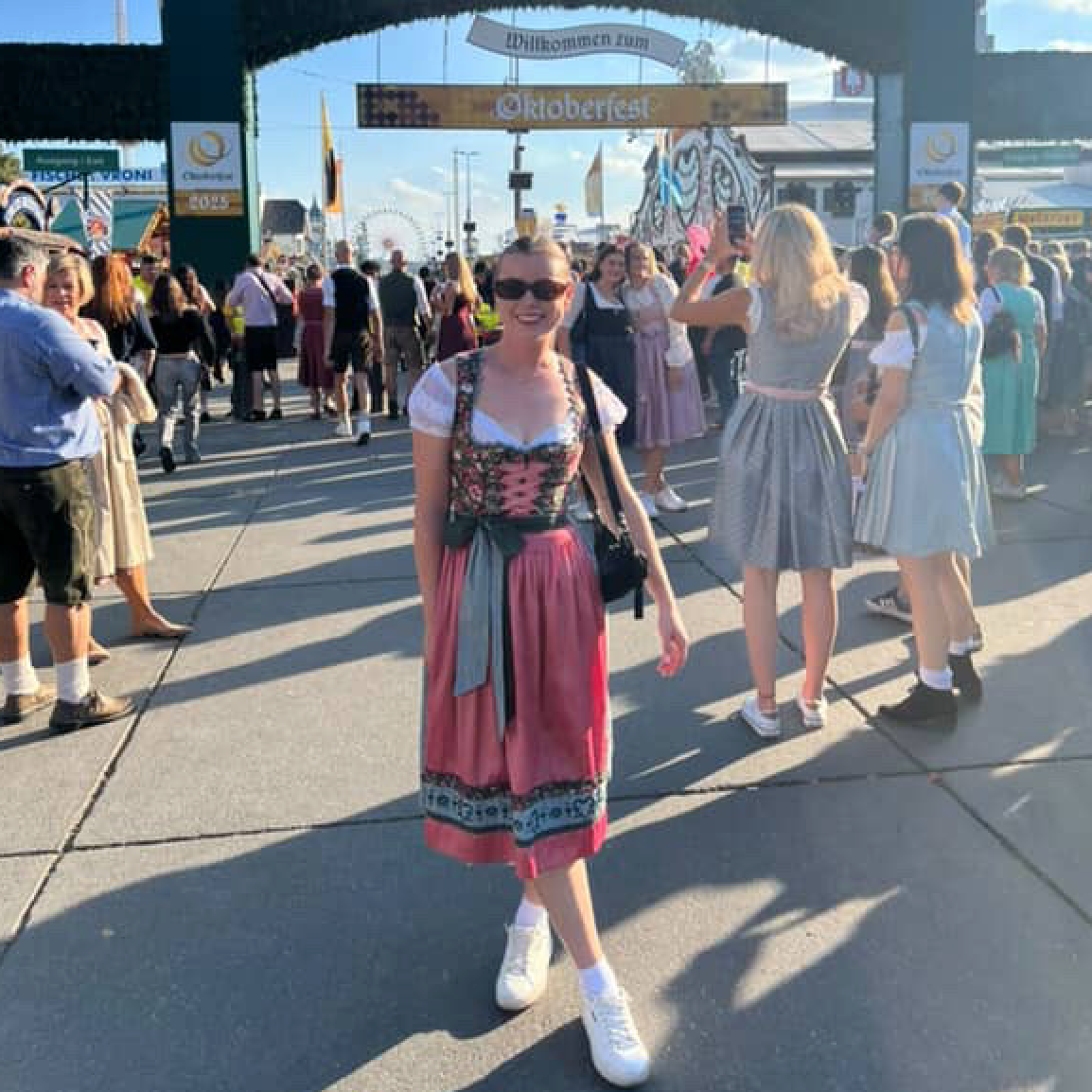 A woman wearing a traditional German dirndl dress our Bestseller Flower Krueger Dirnld in front of the Oktoberfest archway. Dirndl ready to rent on Versatile OTM for Oktoberfest