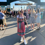 A woman wearing a traditional German dirndl dress our Bestseller Flower Krueger Dirnld in front of the Oktoberfest archway. Dirndl ready to rent on Versatile OTM for Oktoberfest