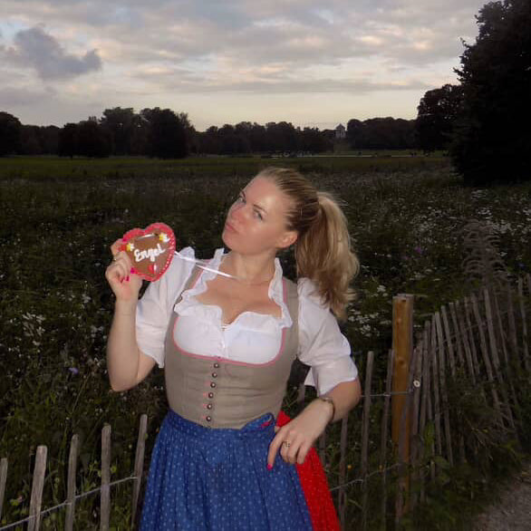 A woman in a traditional German Hammerschmied Dirnd in a crowded Oktoberfest Tent. The dirndl has a white blouse, a beige embroidered bodice with pink trim and brown buttons, and a blue polka-dot skirt, The Hammerschmied Dress is ready to rent on Versatile OTM for Oktoberfest 2026