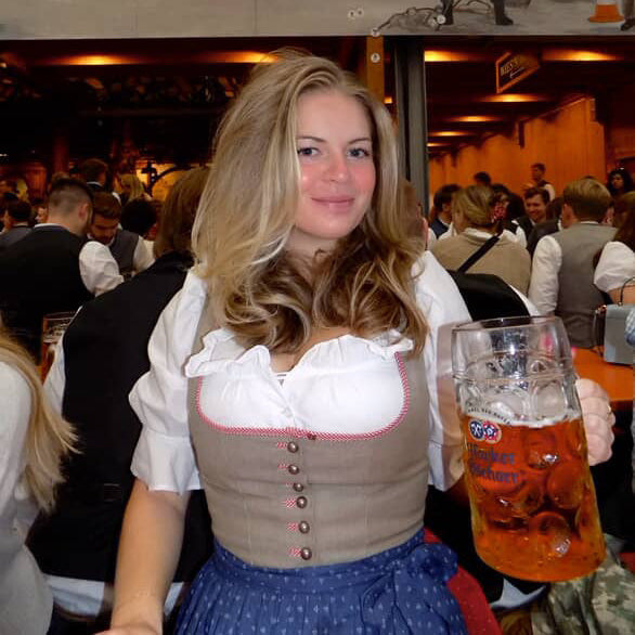 A woman in a traditional German Hammerschmied Dirnd in a crowded Oktoberfest Tent. The dirndl has a white blouse, a beige embroidered bodice with pink trim and brown buttons, and a blue polka-dot skirt, The Hammerschmied Dress is ready to rent on Versatile OTM for Oktoberfest 2026
