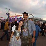 A couple posing in traditional German Dirndl at Oktoberfest. The woman wears a white and silver traditional Dirndl with a heart-shaped Gingerbread Cookie, and the man wears a blue checkered shirt, brown lederhosen. For rental on Versatile OTM website