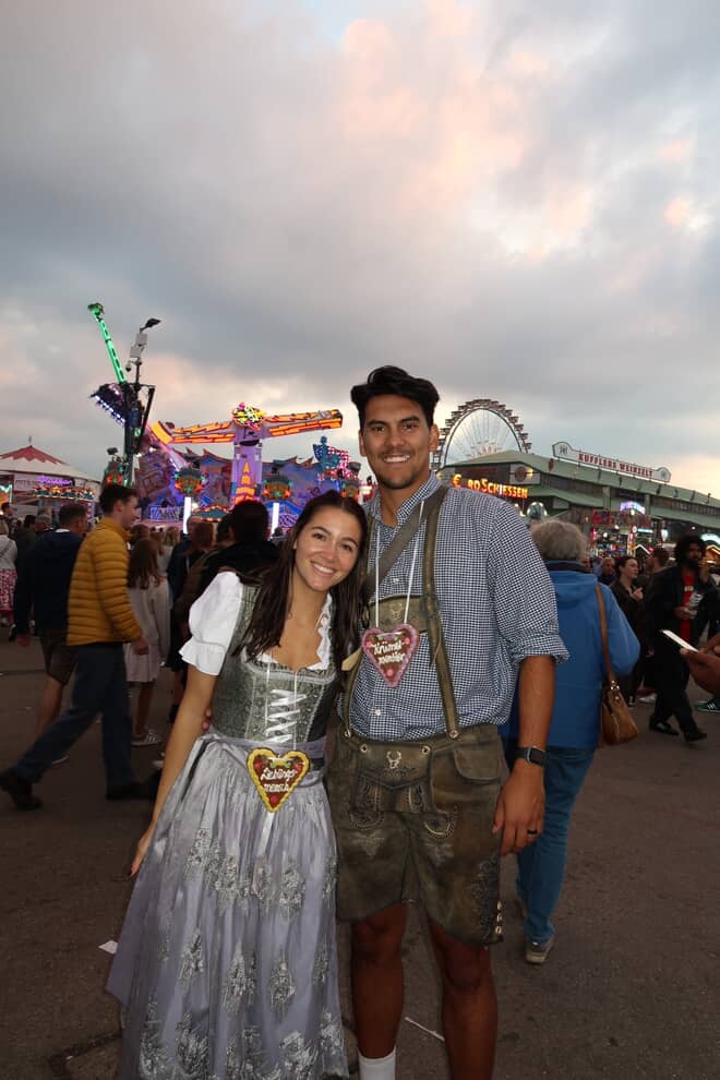 A couple posing in traditional German Dirndl at Oktoberfest. The woman wears a white and silver traditional Dirndl with a heart-shaped Gingerbread Cookie, and the man wears a blue checkered shirt, brown lederhosen. For rental on Versatile OTM website