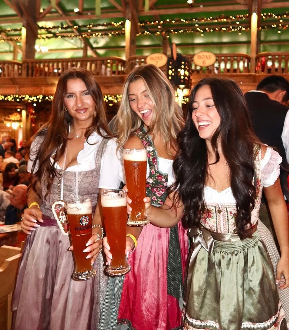 Three women in traditional German Dirndl at Oktoberfest in an Oktoberfest Tent holding beer mugs in a festive setting.