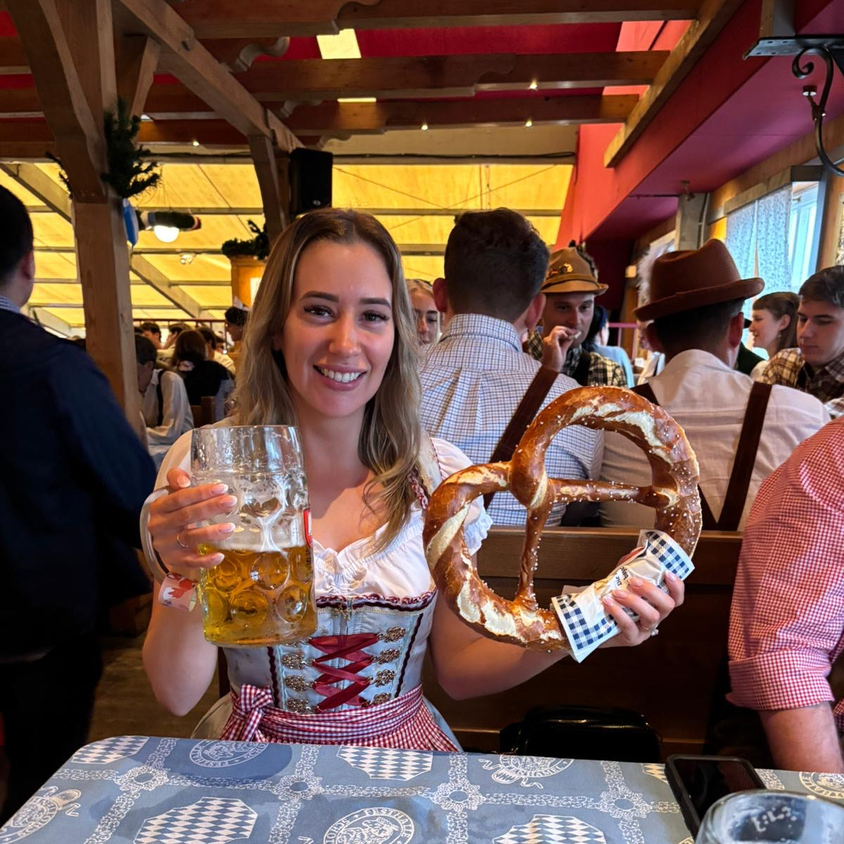 Traditional German Dirndl from Krüger Dirndl for rent on Versatile OTM for Oktoberfest. Woman sitting in a Beer Tent with Pretzel and Beer wearing a rented Dirndl from Versatile OTM 