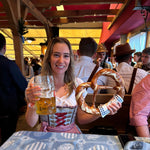 Traditional German Dirndl from Krüger Dirndl for rent on Versatile OTM for Oktoberfest. Woman sitting in a Beer Tent with Pretzel and Beer wearing a rented Dirndl from Versatile OTM 