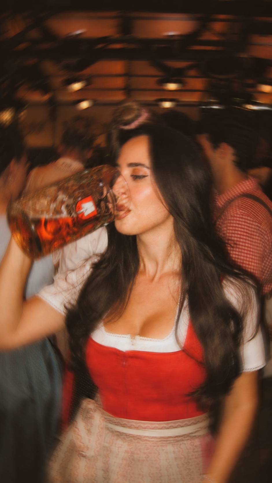 A woman in a red and white traditional outfit drinking from a large beer mug in a beer tent at Oktoberfest 