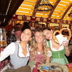 Three women smiling and posing together inside a crowded traditional beer hall, wearing German dirndl outfits. One holds a large beer mug, another holds a pretzel, and the third wears a green dirndl. All Dirndls ready to rent on Versatile OTM 