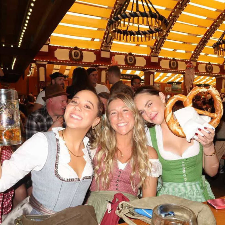 Three women smiling and posing together inside a crowded traditional beer hall, wearing German dirndl outfits. One holds a large beer mug, another holds a pretzel, and the third wears a green dirndl. All Dirndls ready to rent on Versatile OTM 
