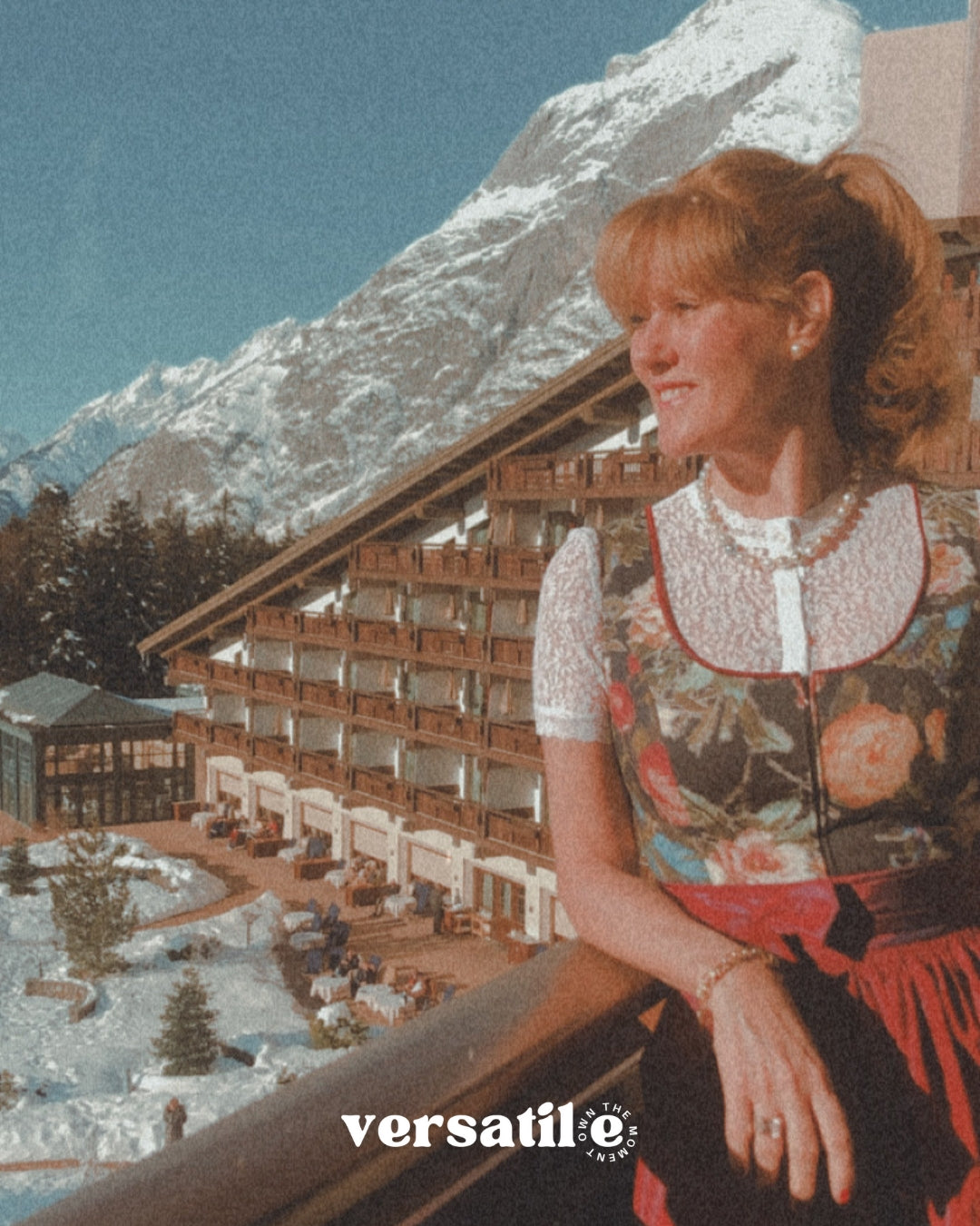 Woman in traditional dress sitting in front of a mountain resort building with 'versatile' branding. Ready to rent via Versatile Versatile Collection Dirndl Celine