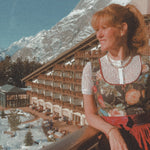 Woman in traditional dress sitting in front of a mountain resort building with 'versatile' branding. Ready to rent via Versatile Versatile Collection Dirndl Celine