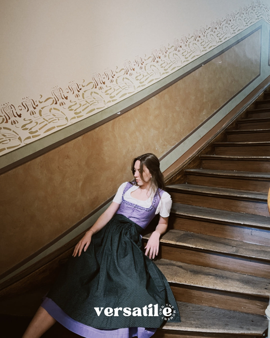 Woman in a traditional outfit sitting on a staircase with 'versatile' branding. Versatile Emilia Dirndl ready to rent 