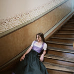 Woman in a traditional outfit sitting on a staircase with 'versatile' branding. Versatile Emilia Dirndl ready to rent 