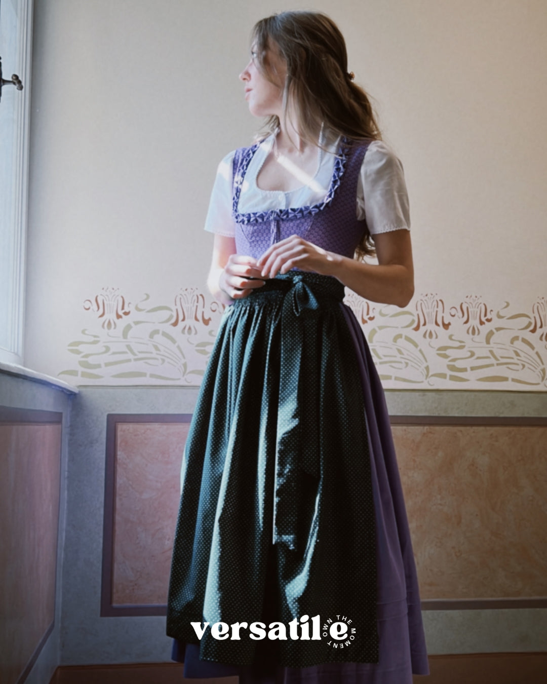 Woman wearing a traditional outfit with a purple top and green skirt in a room with decorative wallpaper. Versatile Emilia Dirndl ready to rent 