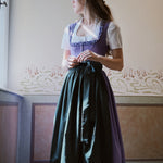 Woman wearing a traditional outfit with a purple top and green skirt in a room with decorative wallpaper. Versatile Emilia Dirndl ready to rent 
