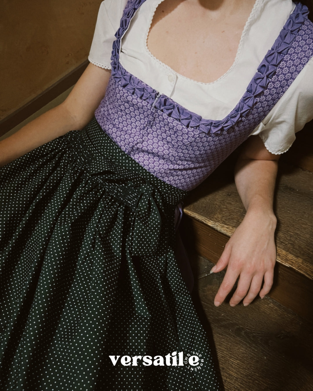 Person wearing a traditional outfit with a purple top and black polka dot skirt, sitting on a wooden floor. Versatile Emilia Dirndl ready to rent 