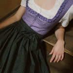 Person wearing a traditional outfit with a purple top and black polka dot skirt, sitting on a wooden floor. Versatile Emilia Dirndl ready to rent 