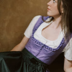 Woman wearing a purple green Dirndl Dress top over a white shirt with a brown background. Versatile Emilia Dirndl ready to rent 