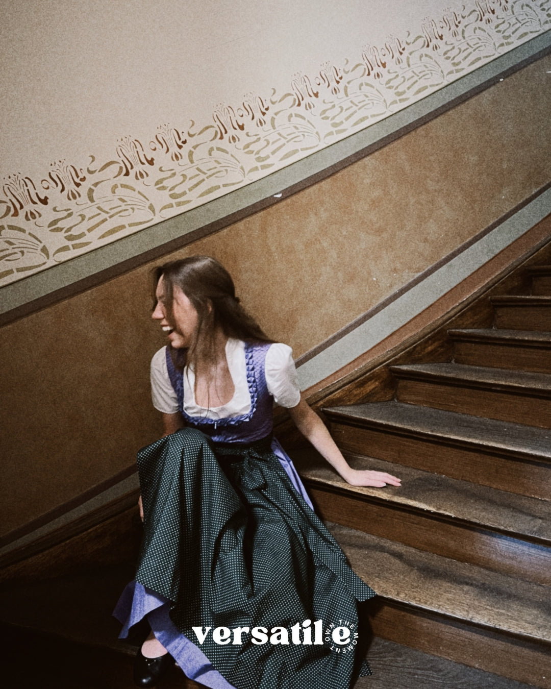 Woman in a vintage Dirndl Dress sitting on a staircase with 'versatile' branding. Versatile Emilia Dirndl ready to rent 
