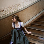 Woman in a vintage Dirndl Dress sitting on a staircase with 'versatile' branding. Versatile Emilia Dirndl ready to rent 