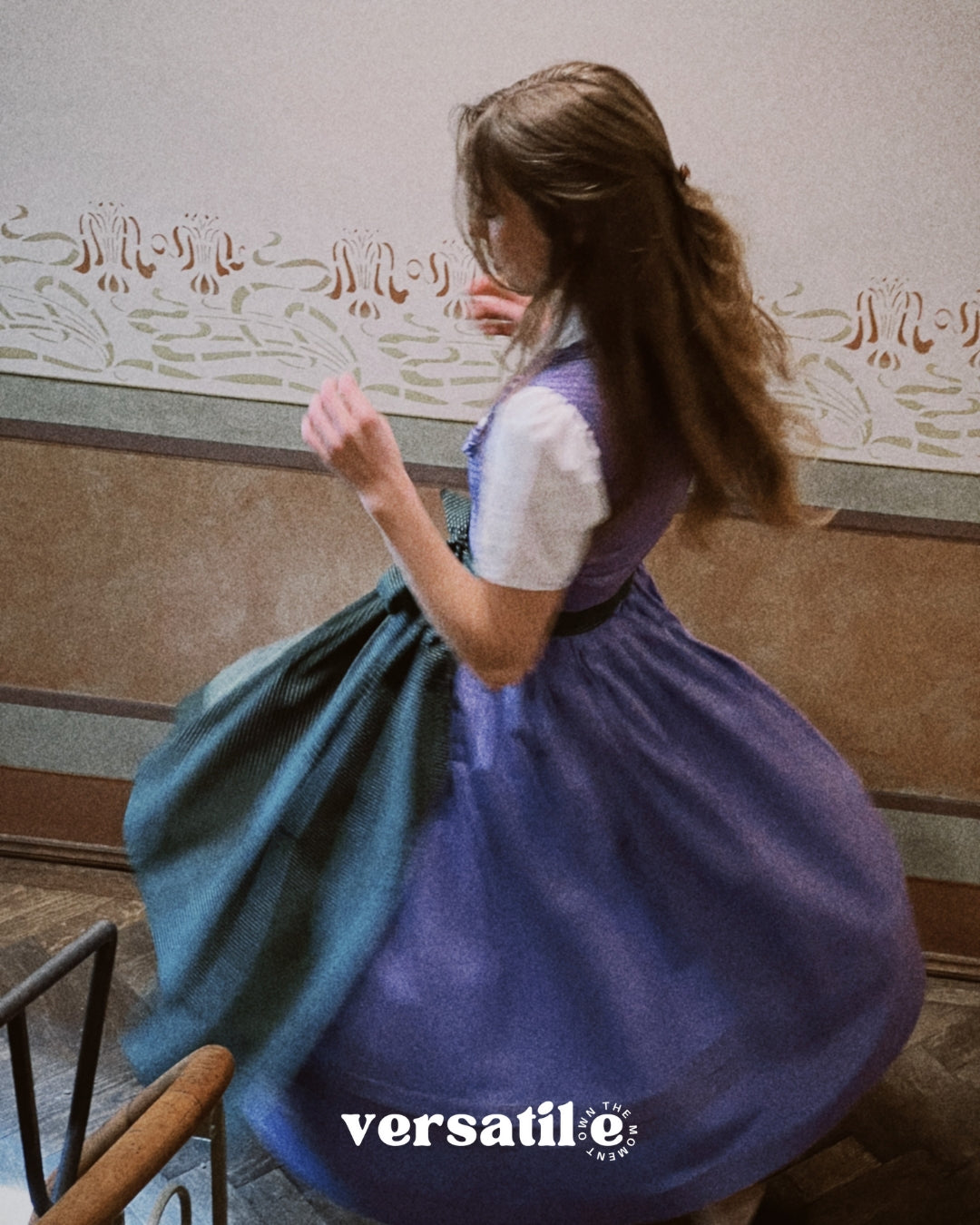 Person in a purple Dirndl dress with a white blouse, sitting on a bench with a decorative wall in the background. Versatile Emilia Dirndl ready to rent 