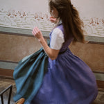 Person in a purple Dirndl dress with a white blouse, sitting on a bench with a decorative wall in the background. Versatile Emilia Dirndl ready to rent 