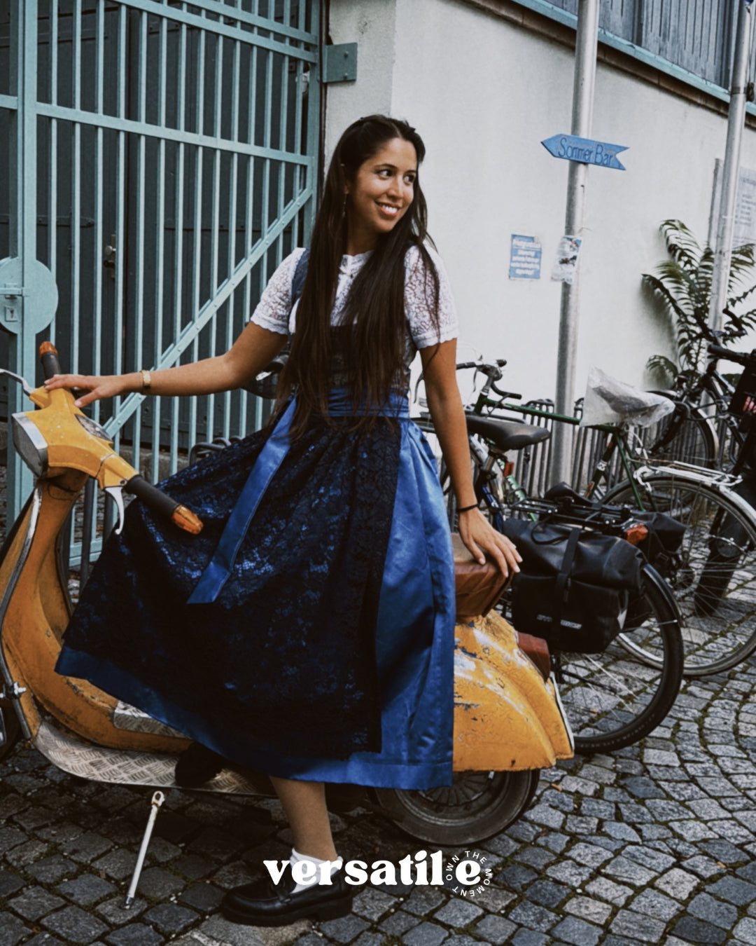 Woman in a blue dress Dirndl Pia from the versatile collection standing next to a scooter with bicycles in the background, featuring the brand 'versatile'. Ready for Oktoberfest