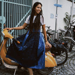 Woman in a blue dress Dirndl Pia from the versatile collection standing next to a scooter with bicycles in the background, featuring the brand 'versatile'. Ready for Oktoberfest