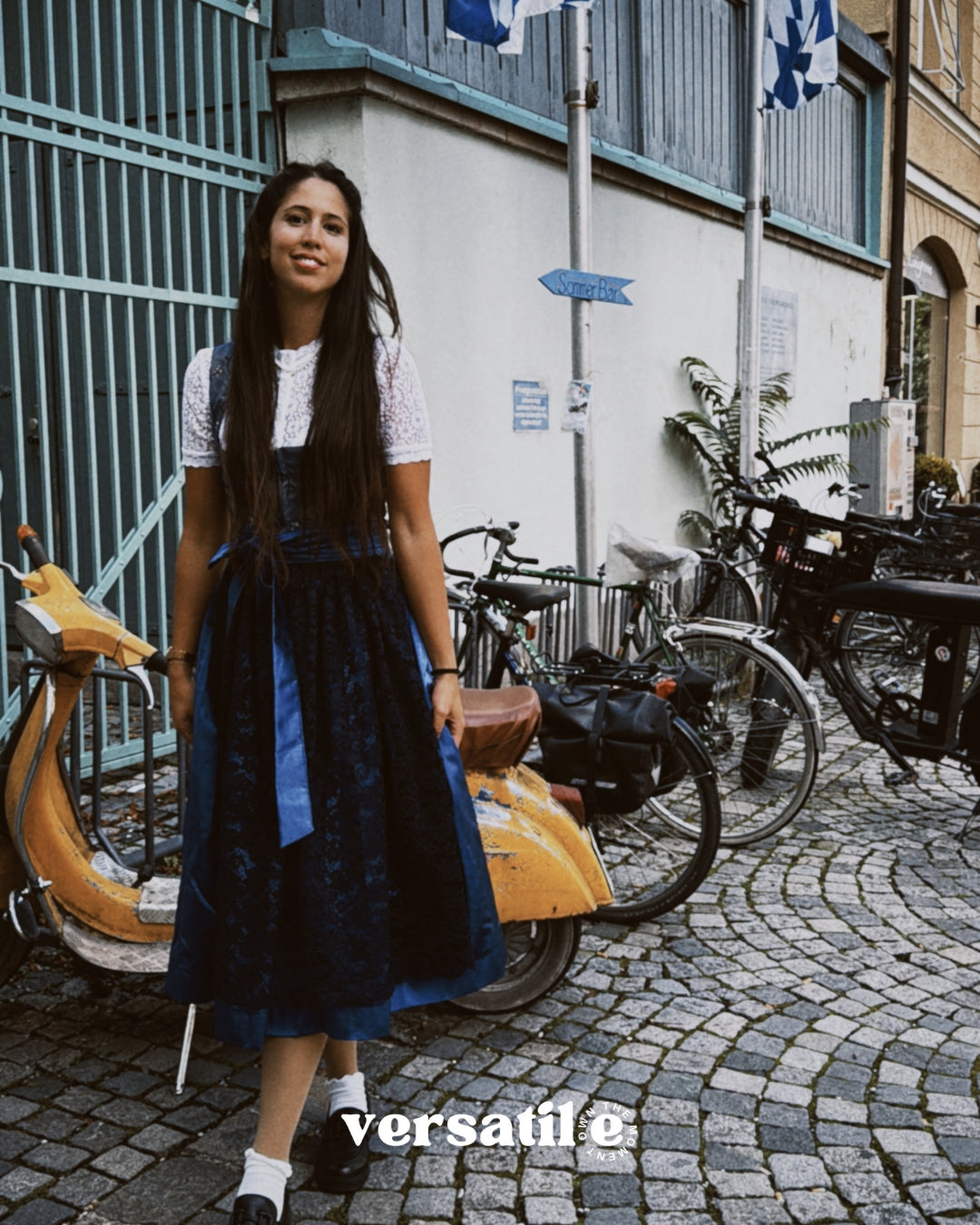 Woman in a blue dress standing on a street with bicycles and buildings in the background, featuring the brand 'versatile'.