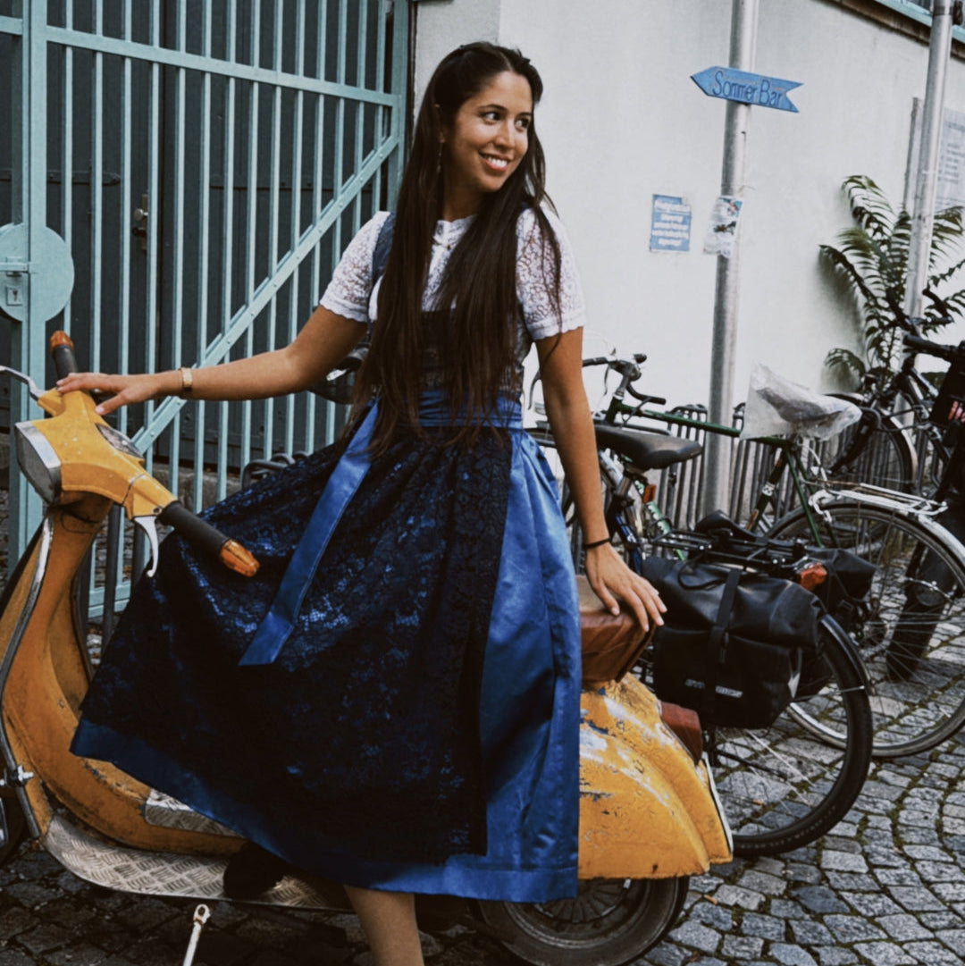 Woman in a blue dress Dirndl Pia from the versatile collection standing next to a scooter with bicycles in the background, featuring the brand 'versatile'. Ready for Oktoberfest