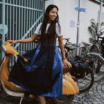 Woman in a blue dress Dirndl Pia from the versatile collection standing next to a scooter with bicycles in the background, featuring the brand 'versatile'. Ready for Oktoberfest